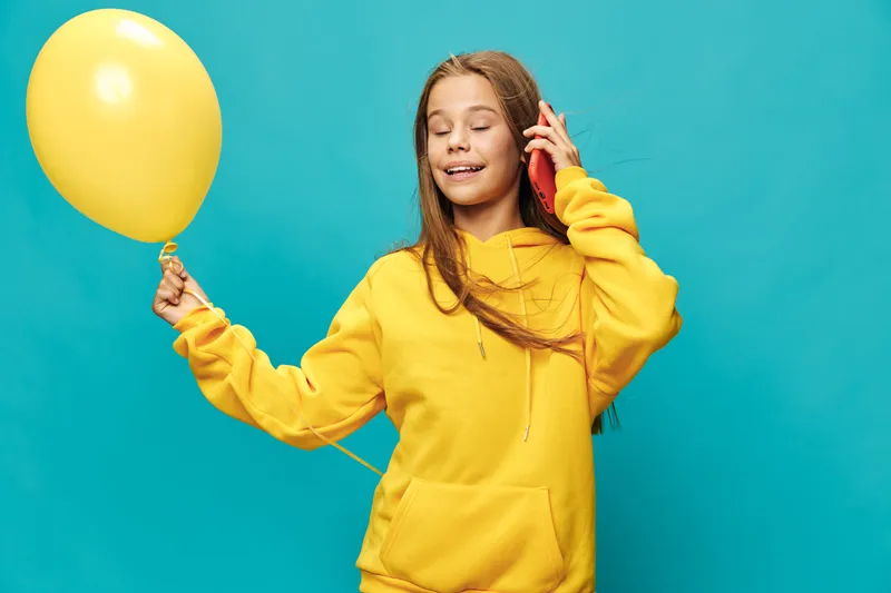 Girl in yellow hoodie holds balloon and talks on phone with closed eyes smiling, isolated on blue background for celebration, happiness and communication concept.