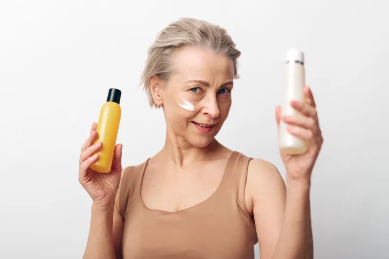 Mature woman with short hair holds two skincare bottles and applies cream on cheek. Concept of beauty routine, anti aging, self care, and skin hydration in studio with white background.