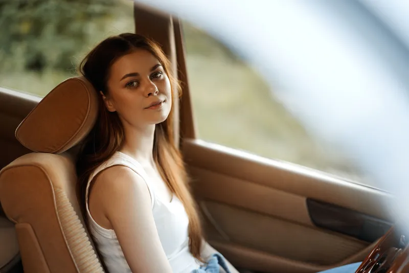 Young woman with long brown hair sitting in a car, wearing a casual outfit and smiling softly, surrounded by a natural outdoor background.