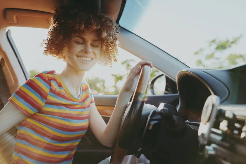 Smiling young woman with curly hair sits behind the steering wheel in a sunlit car, enjoying a carefree moment during a casual road trip.