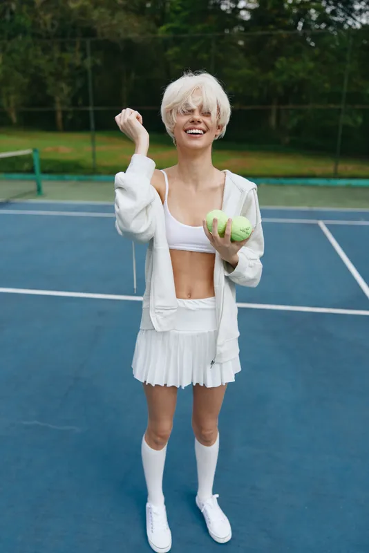 Joyful young woman in sporty attire on a tennis court, wearing a white crop top and pleated skirt, representing vibrant energy, fitness, and summer fun