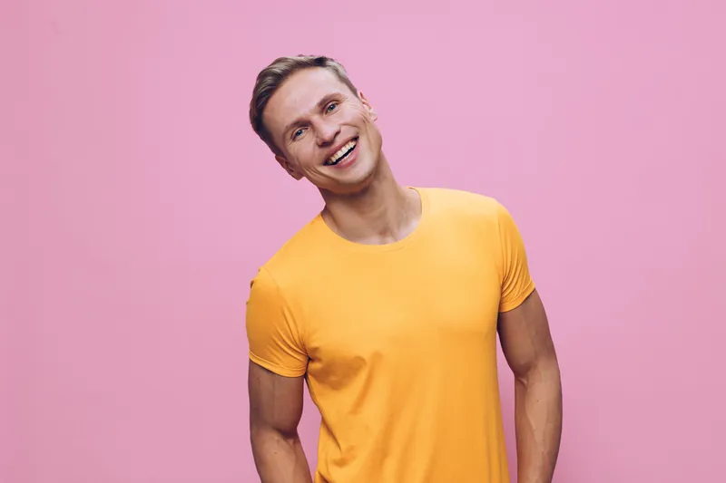 Smiling happy young caucasian man wearing orange t-shirt standing isolated on pink background cheerful friendly emotion concept