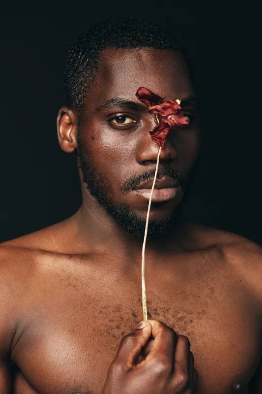 Creative conceptual portrait of African man with dark skin holding dried flower in front of face on black background. Artistic visual style with focus on expression and texture.