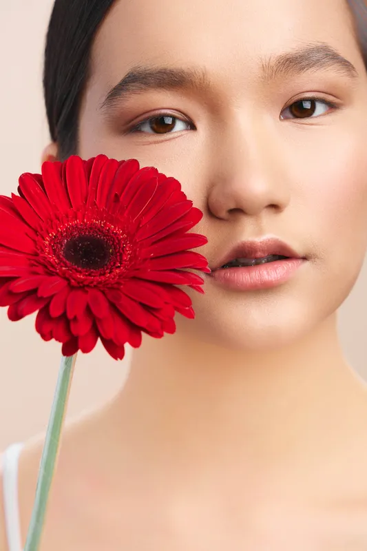 Asian woman poses with a vivid red flower close to her face, soft lighting highlights smooth skin and a serene expression, creating an elegant, contemporary portrait.