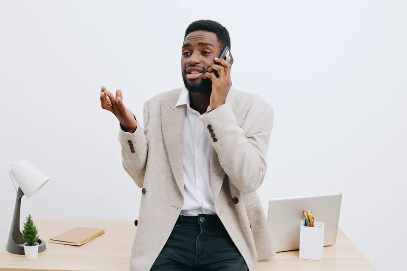 Professional man talking on mobile phone while gesturing with his hand, standing at modern office desk with laptop, notepad, and decorative plant, expressing engagement and communication in a business
