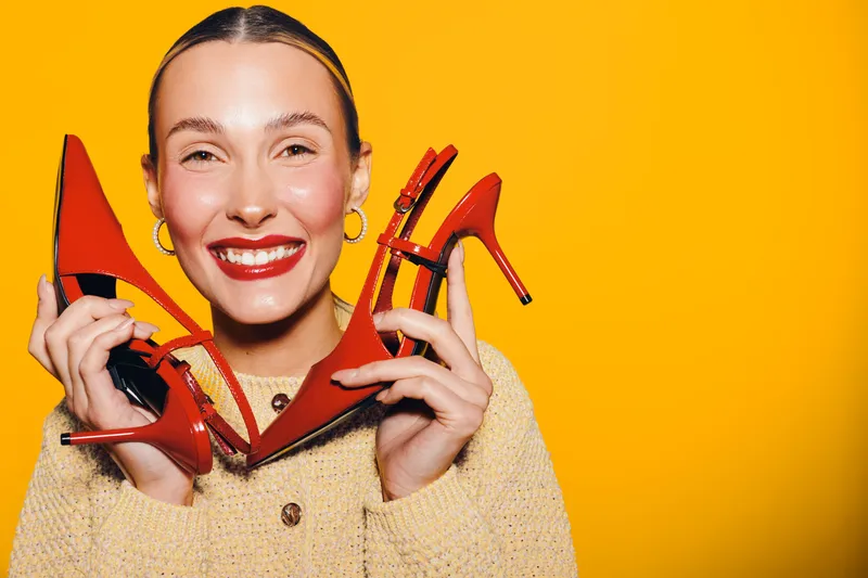 Smiling woman with red lipstick holds red high heel shoes near face on yellow background. Concept of fashion, style, beauty, and happiness with bright studio lighting and joyful mood.