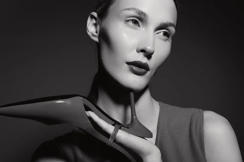 Close-up portrait of a fair skinned white woman holding a high heel shoe near her face, posing in dramatic black and white lighting. Concept of fashion and elegance.