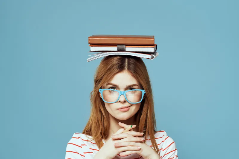 Студентка держит книги на голове на фоне синего цвета, что символизирует научное образование