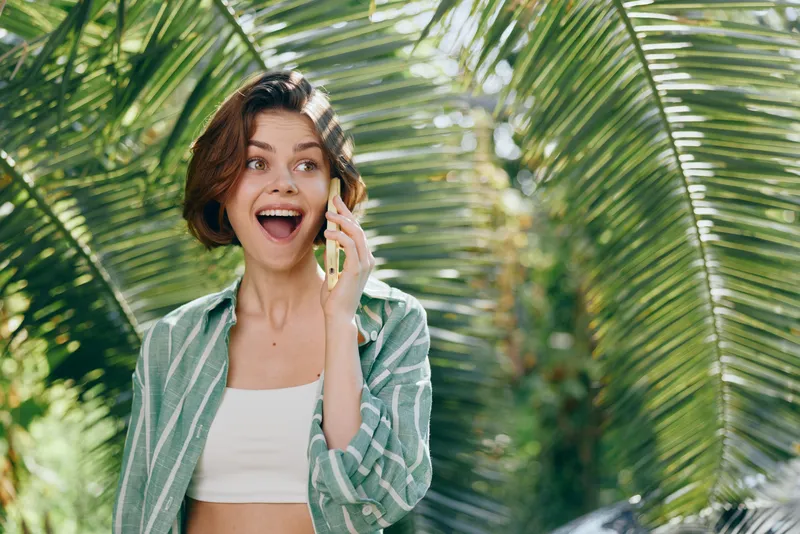 A smiling woman on a sunny day talks on a phone, framed by lush green palm leaves in a bright outdoor setting.