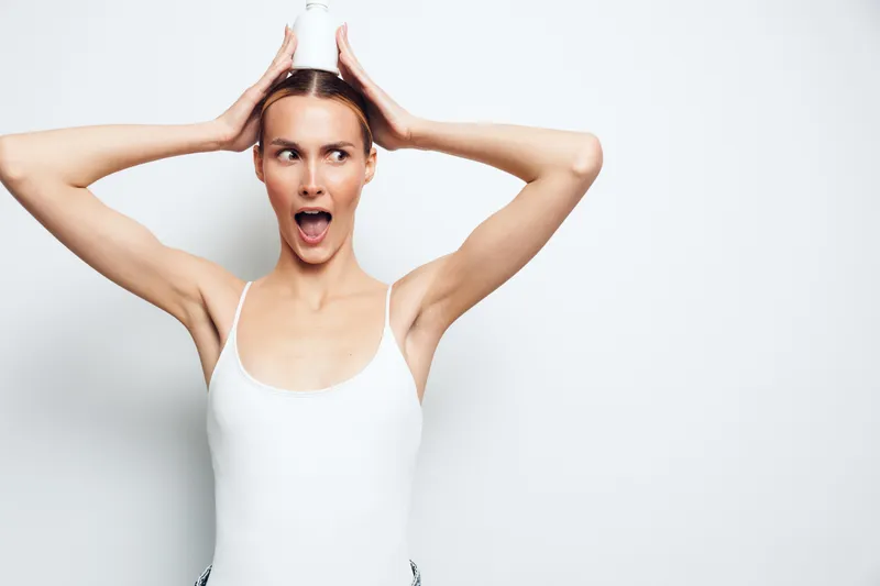 Young woman in white tank top holding a small white bottle on her head, surprised expression, minimalistic background, casual pose, bright lighting, studio shot