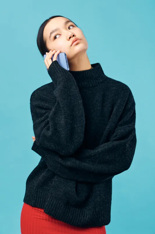 Young woman in black sweater talking on smartphone against blue background looking away thoughtfully. Casual clothing, communication, mobile phone and modern technology concept portrait.