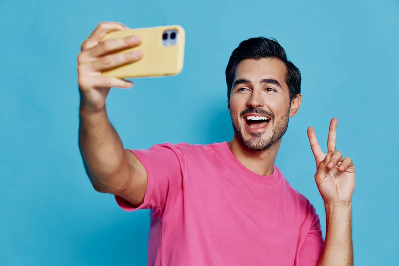 Full body smiling happy cheerful young man takes selfie using mobile phone, posing with peace sign against blue background. Social media lifestyle concept.