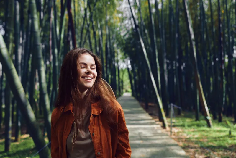 Woman smile forest path happy portrait nature walking, young female outdoors on a wooded trail in a casual jacket, eyes closed and smiling, peaceful relaxed moment in green scenic environment.