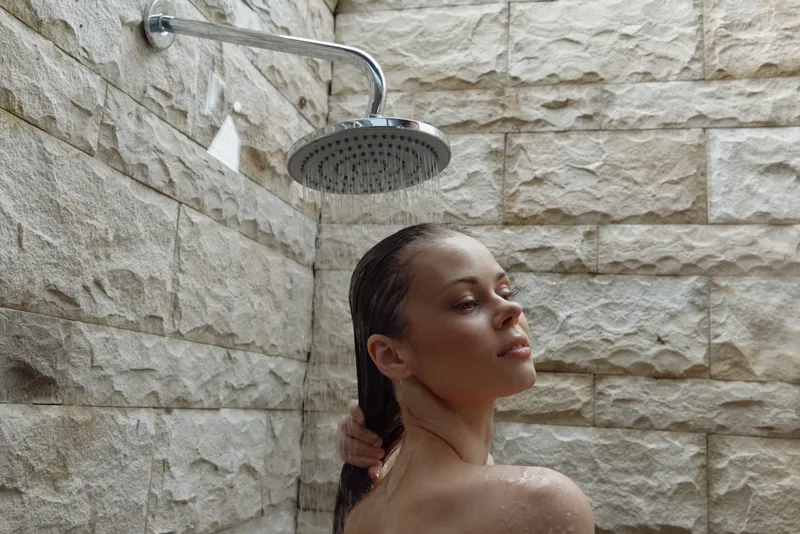 Young woman relaxing in outdoor shower with stone wall background, natural light, wet hair, and peaceful expression in warm, calm atmosphere.