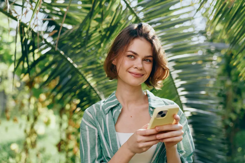 Cheerful woman outdoors, holding smartphone, relaxed pose, sunny day, lush greenery, casual attire, warm smile, ready for a moment