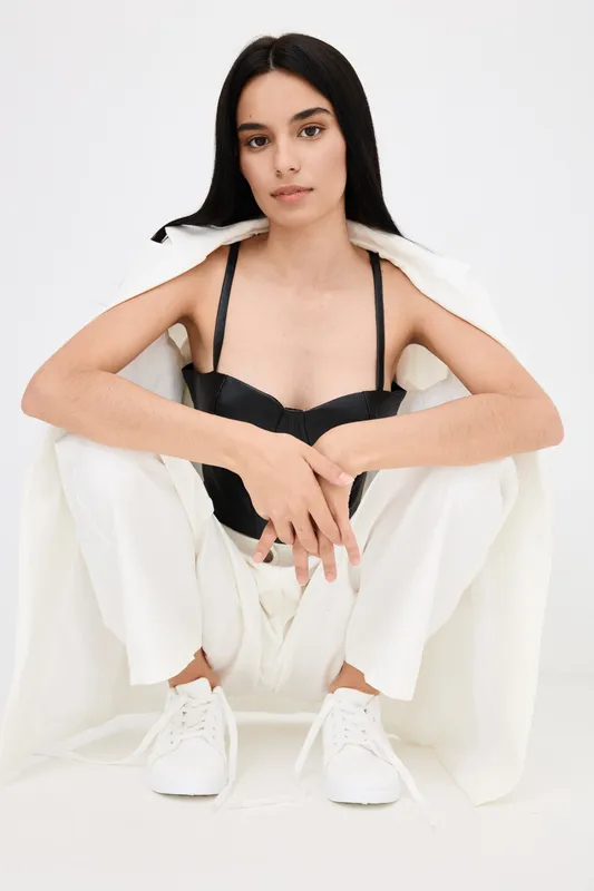 Young woman sitting wearing white pants and sneakers with a black top and white jacket draped over her shoulders in a minimal studio setting.