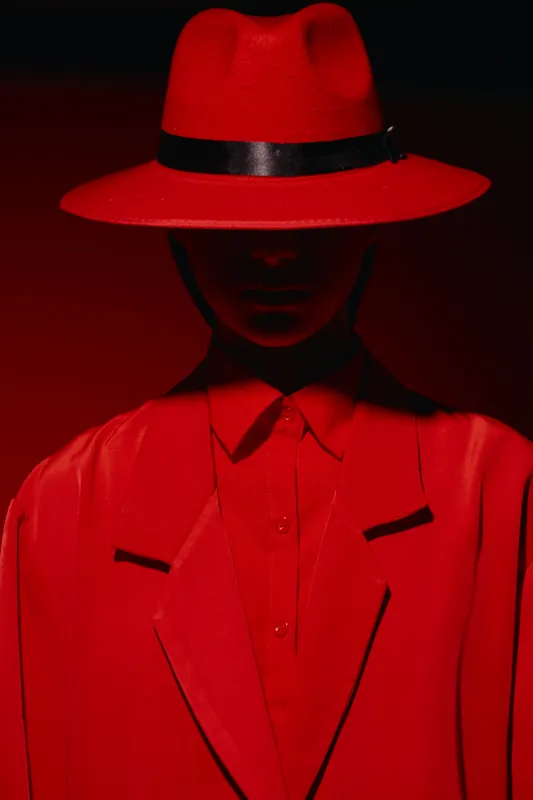 Stylish model wears striking red fedora hat and matching suit against a deep red background with dramatic shadows. Concept for fashion, mystery, and creative beauty portrait.