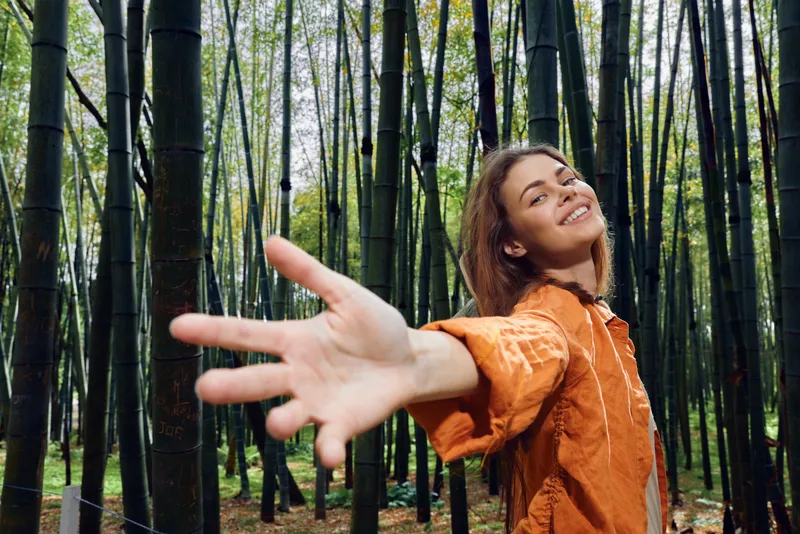 Woman smile in bamboo forest reaching hand toward camera in outdoor nature portrait. Joyful young traveler in orange jacket exploring serene grove with playful connection.