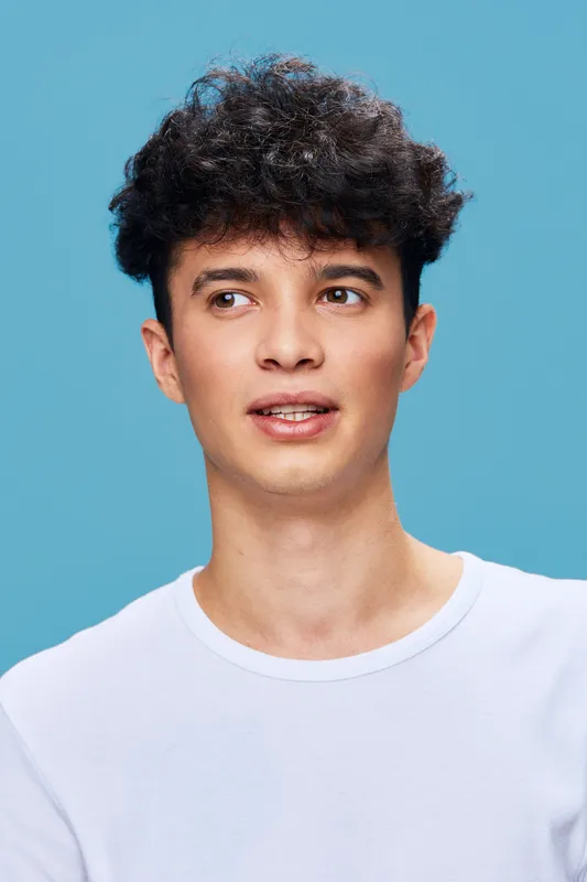 Young man with curly hair wearing a white t-shirt, looking away thoughtfully against a solid light blue background, casual style and natural expression.
