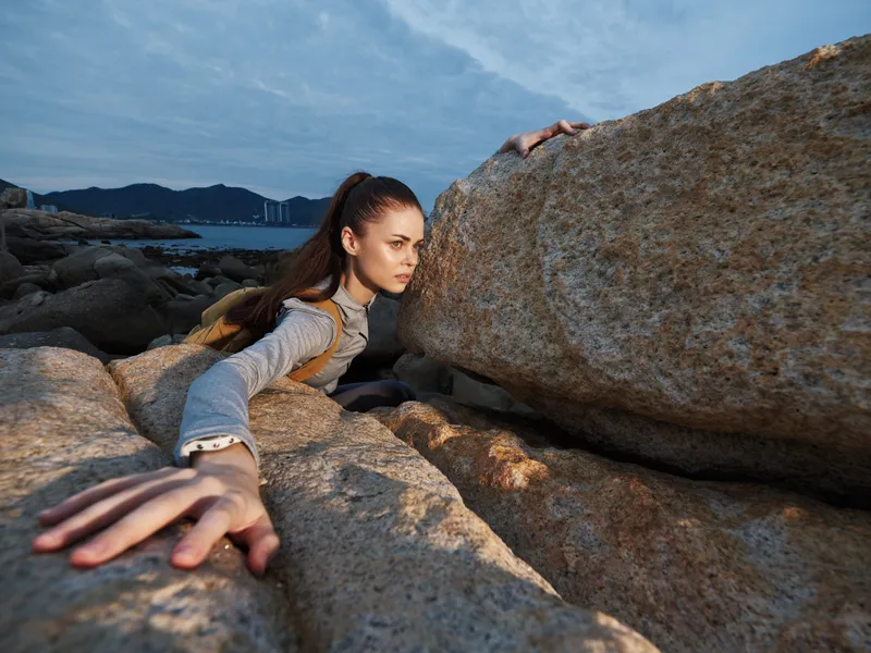 Young woman climbing between rocks at the beach during sunset, showcasing determination and strength in an outdoor adventure setting