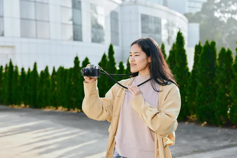 Camera worn by a woman outdoors, a photographer holds a handheld camera with strap, casual jacket, calm smile, sunlit park and modern building backdrop, lifestyle image for travel, hobby, and street