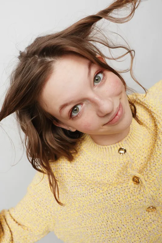 Young woman with green eyes and freckles smiles gently wearing a yellow knitted sweater. Close-up portrait with natural hair and soft lighting on a neutral background.