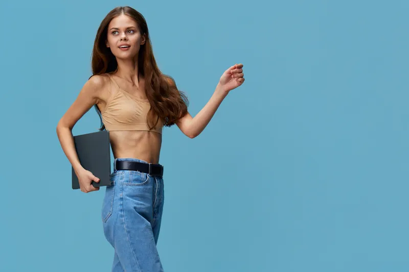 young woman holding laptop confidently wearing casual beige top and blue jeans posing against blue background with long hair and charming smile