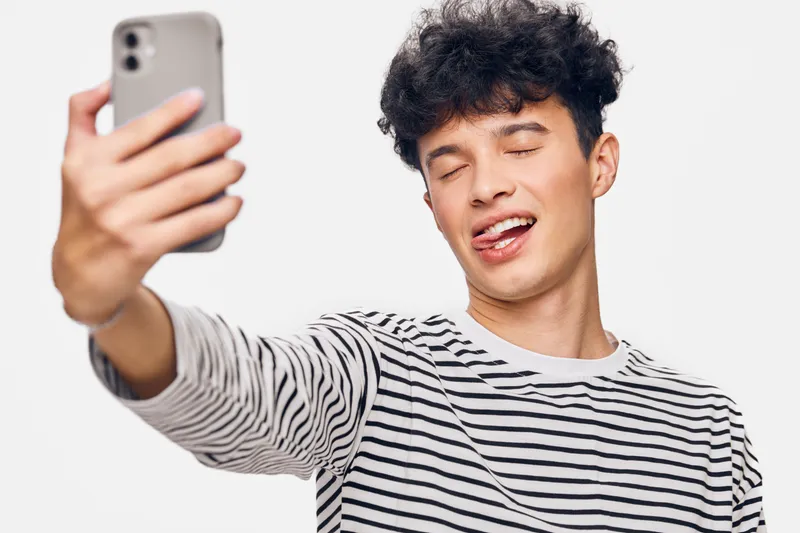 young man taking selfie with smartphone, wearing striped shirt, playful expression, white background, casual style, modern technology