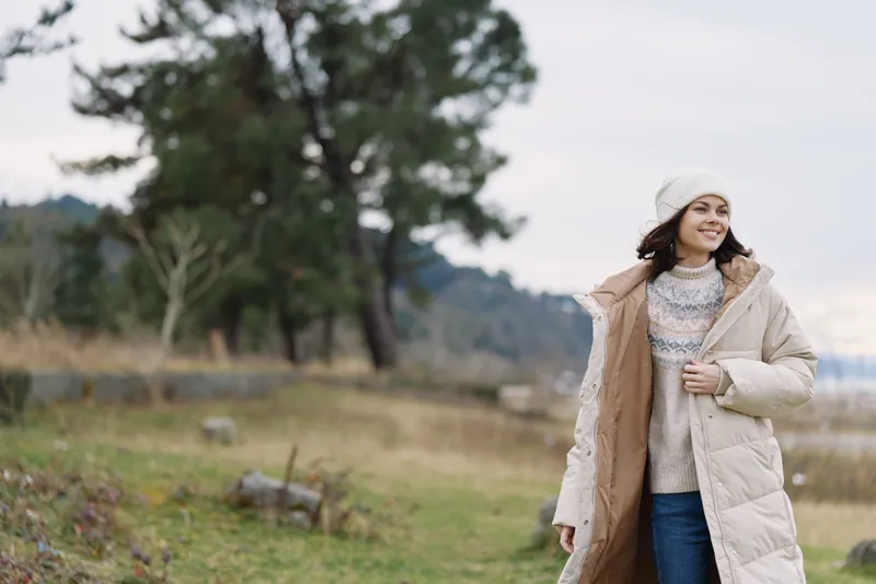 Woman outdoors in a long beige winter coat and cozy knit sweater, walking along a lakeside path with trees and grass, capturing a calm seasonal mood and effortless urban style.