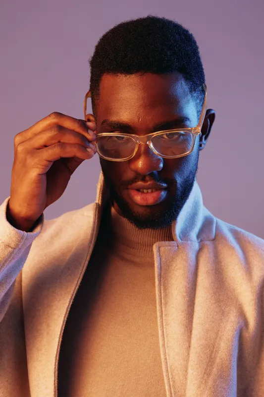 Confident young Black man wearing glasses and a stylish coat, posing with a captivating expression against a soft colored background