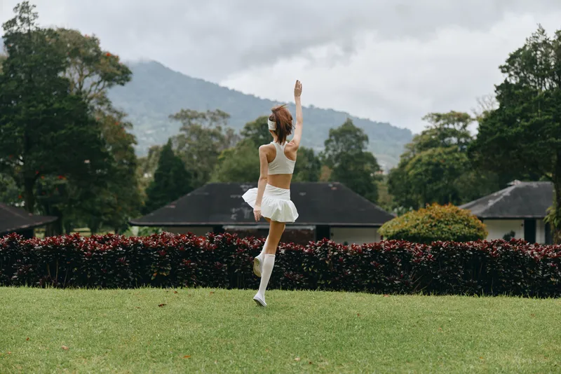 A young woman in a white sporty outfit dancing joyfully in a vibrant green park, showcasing her happiness against a scenic mountain backdrop