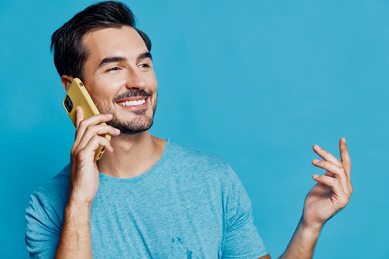 Smiling man talking on mobile phone wearing casual blue t-shirt with a cheerful expression against a vibrant blue background. Modern lifestyle concept.