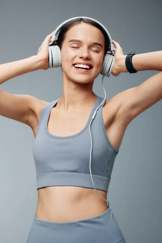 Fitness woman in a sporty outfit enjoying music through headphones during a workout, smiling with energy and focus in a clean studio backdrop.