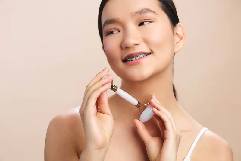 Asian woman wearing a gentle smile holds a skincare device near her cheek in a soft studio setting, conveying a calm, modern selfcare moment and healthy radiant glow.