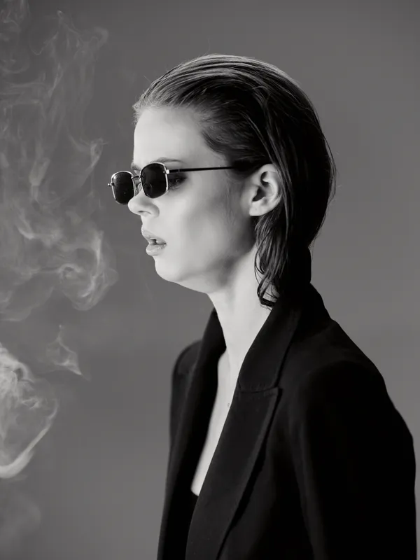Black and white portrait of a woman wearing sunglasses smoking with a confident expression in a dark blazer.