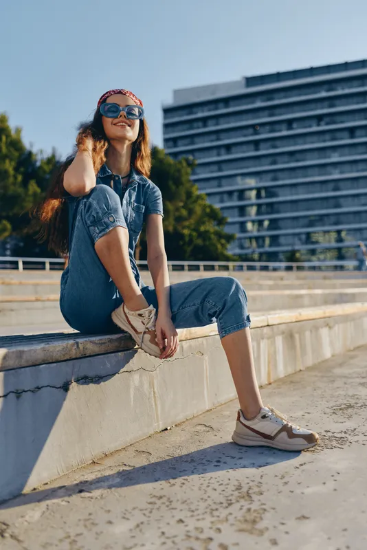 smiling woman in denim jumpsuit wearing sunglasses and sneakers sitting outdoors on concrete steps under bright sunlight in urban environment