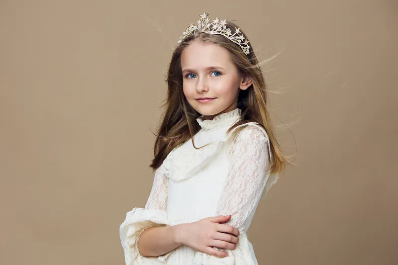 Young girl in a white princess dress with a crown, smiling confidently against a beige background, embodying innocence and charm Perfect for children s fashion themes
