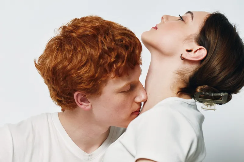 Couple, intimacy, and affection with young redhead man kissing woman neck in studio on white background. Passion, love, romance, and connection in relationship between two people.
