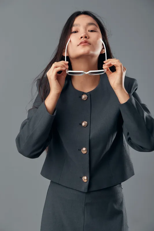 Woman in business suit adjusts eyeglasses in professional studio setting, confident young female model with long dark hair and formal outfit on gray background for corporate concept.