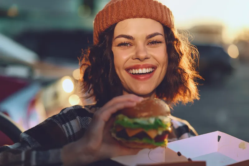 Woman smile burger outdoor candid shot capturing authenticity and candid lifestyle during golden hour glow, mindful living and emotional storytelling in urban evening street food.