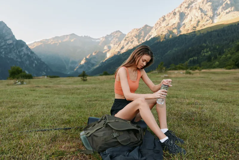 A young woman sits on grass in a scenic valley with towering mountains, stretching after an outdoor workout. The tranquil scene conveys health, focus, and nature.