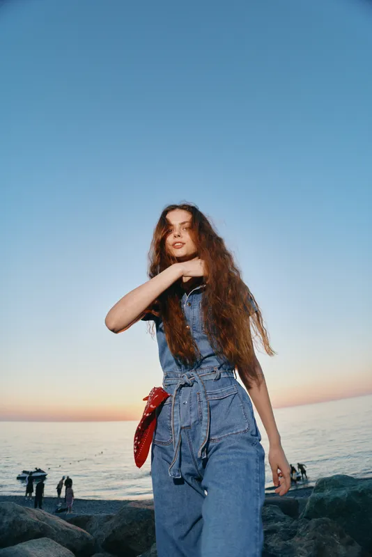 Lifestyle woman in modern boho-western denim street style with long hair stands by the rocky seaside at sunset, film color tone enhancing the relaxed atmosphere.