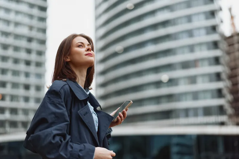 Smiling woman in a navy coat stands outdoors in an urban setting, holding a phone. Clear city backdrop conveys professional lifestyle and modern city vibe.