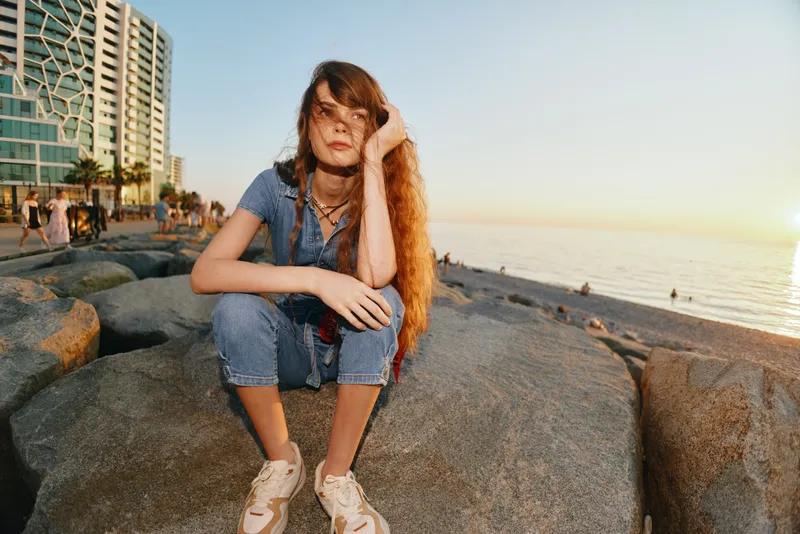 young woman sitting on rocks by the sea shore during golden hour with long hair and casual clothes in coastal urban background