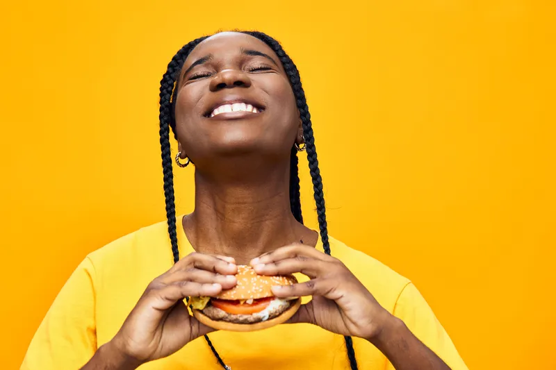 Joyful young Black woman enjoying a hamburger against a vibrant yellow background, showcasing happiness, comfort food, and a cheerful vibe in summer fashion