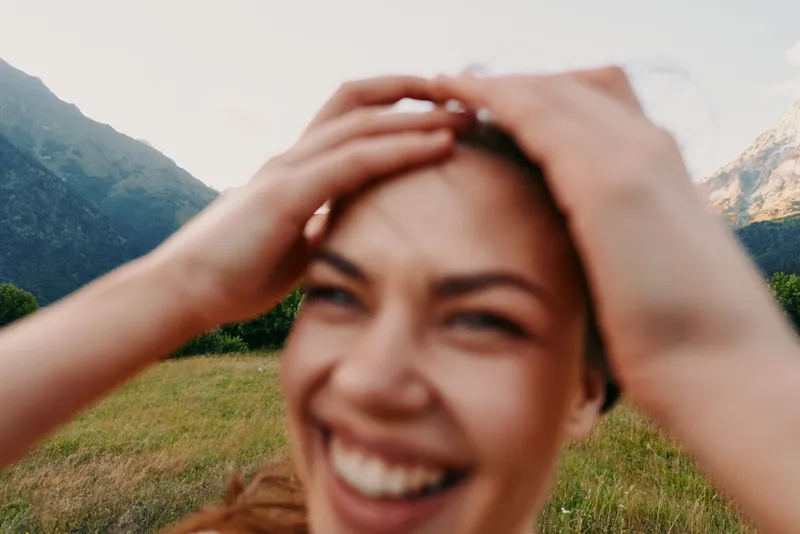 Smiling woman outdoors in a grassy field with mountains in the background, casual lifestyle, wellness vibe, natural light, cheerful candid moment and carefree energy.