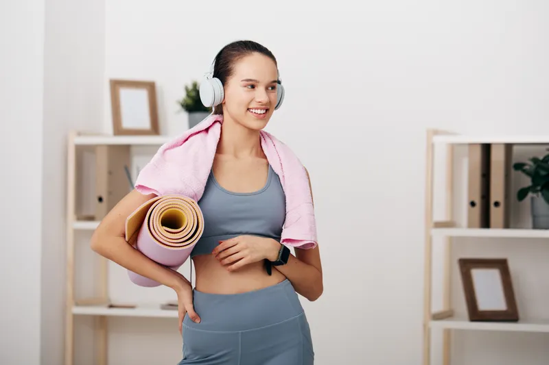 Female fitness enthusiast wearing headphones, carrying a yoga mat and towel over shoulders in a bright indoor studio, ready for a focused workout and training session.