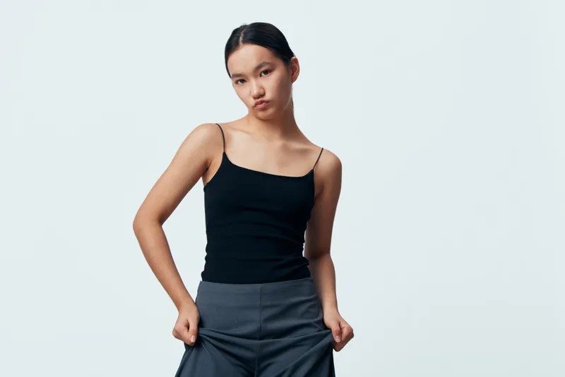 Young woman posing with hands in pockets wearing black tank top and gray pants standing against plain white background. Straight hair, casual style and confident facial expression with minimal makeup.