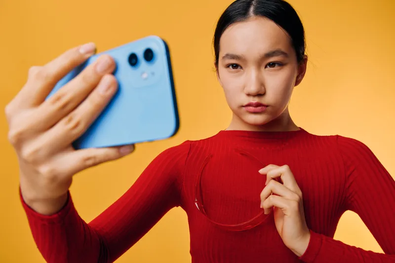 Woman in red top taking selfie with smartphone on yellow background. Young female focused on device for social media content or personal photo. Studio portrait with casual expression and modern
