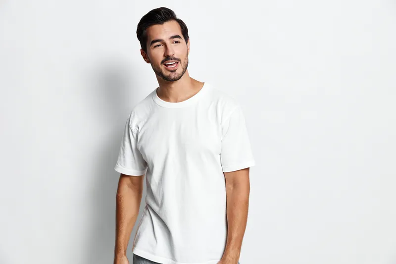 Full body smiling cheerful young man wearing a white t-shirt, standing against a plain light background, looking at the camera with a joyful expression.